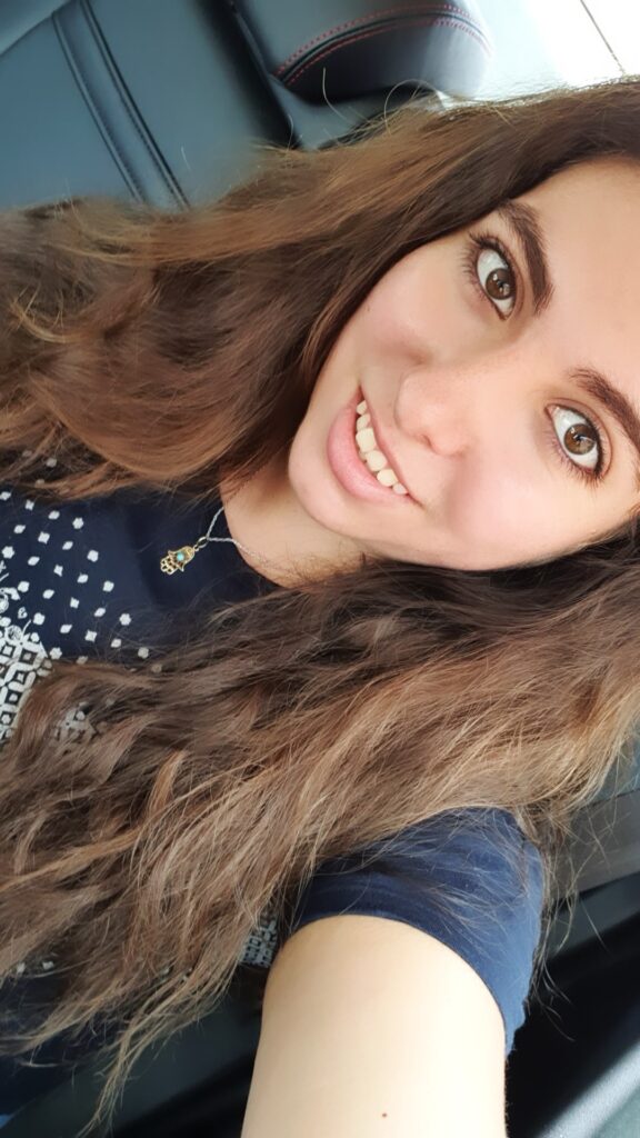 Fotografía de un mujer de 27 años, cabello largo ondulado castaño, ojos castaños. Con una camiseta azúl. Tiene un collar con una hamsa y está sonriendo.