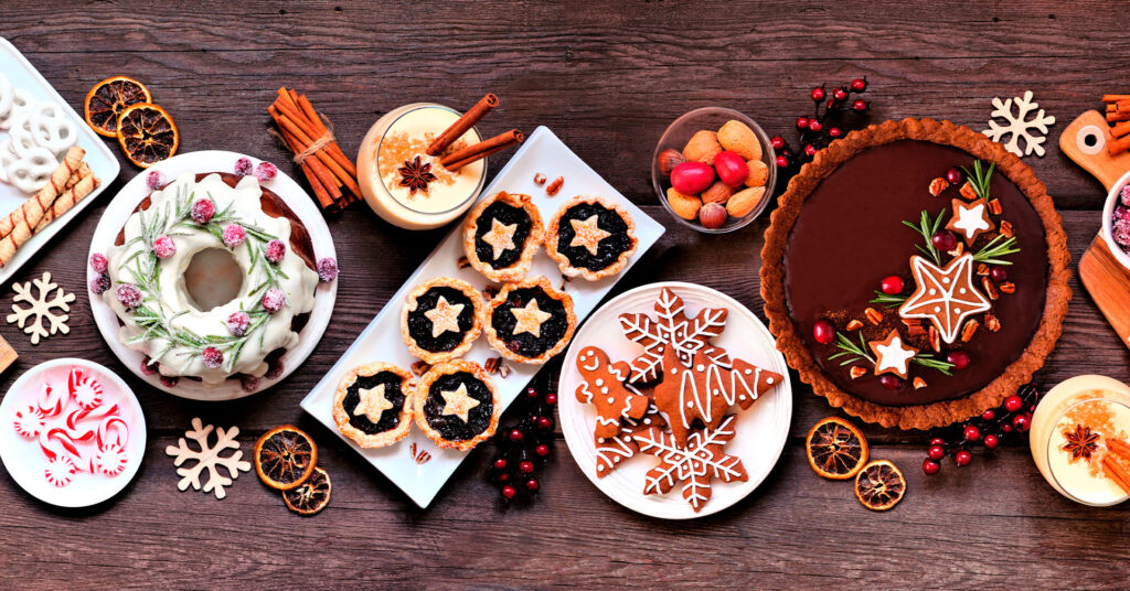 Fotografía con una variedad amplia de postres navideños: pastel de chocolate, tartaletas, caramelo, ponche de huevo, galletas de jengibre y una rosca.