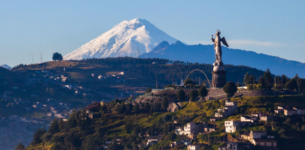 Foto panorámica de Quito donde en un primer plano está el Panecillo, a sus pies están los diferentes barrios que se han construido en la ladera y al fondo está el volcán Cotopaxi, el volcán está nevado.
