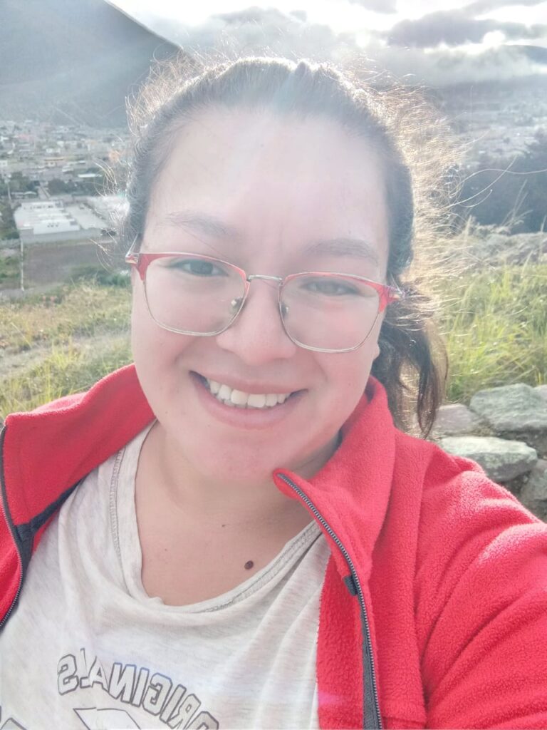 En la imagen se ve una mujer joven sonriendo alegremente al aire libre bajo un sol brillante que ilumina su cabello recogido. Lleva puestos sus lentes de marco rosado, una camiseta clara y un saco de color rojo vivo, con un fondo de montañas verdes y un pueblo.