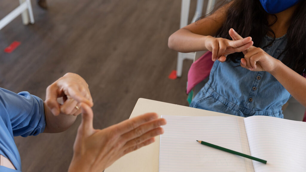 Fotografía inclusiva done la lengua de señas es el puente de comunicación, en la imagen se observa  dos personas sentadas en la mitad una mesa con un cuaderno, la primera persona esta enseñando a la alumna, mientras que la alumna replica lo mismo. 